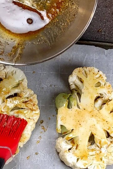silicone pastry brush brushing the top of the cauliflower steaks on a sheet pan with parchment paper